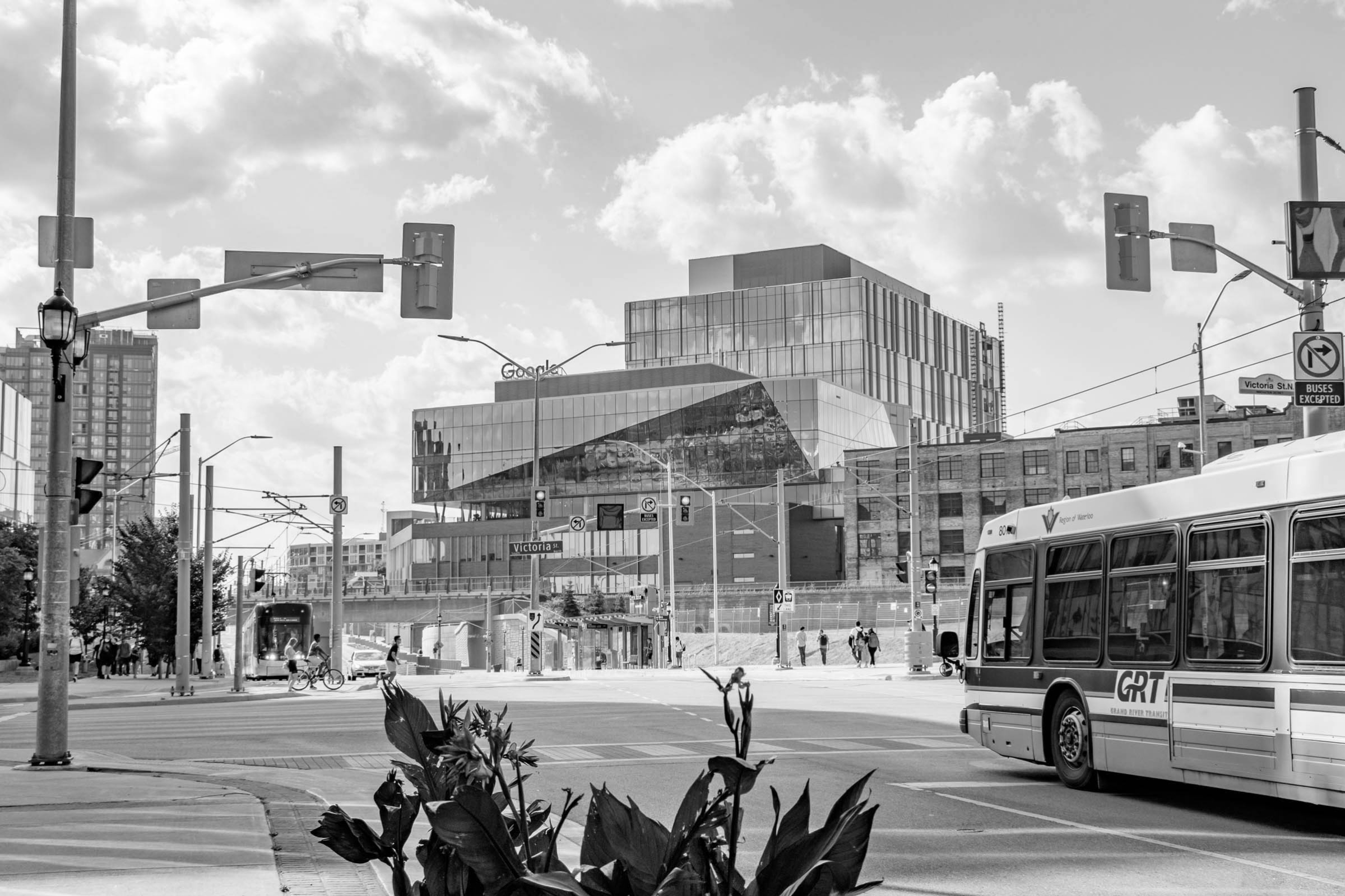 a photo taken at the king st and victoria st intersection looking at the Google building with a GRT bus entering the shot, an LRT Ion train in the background and pedestrians walking and biking