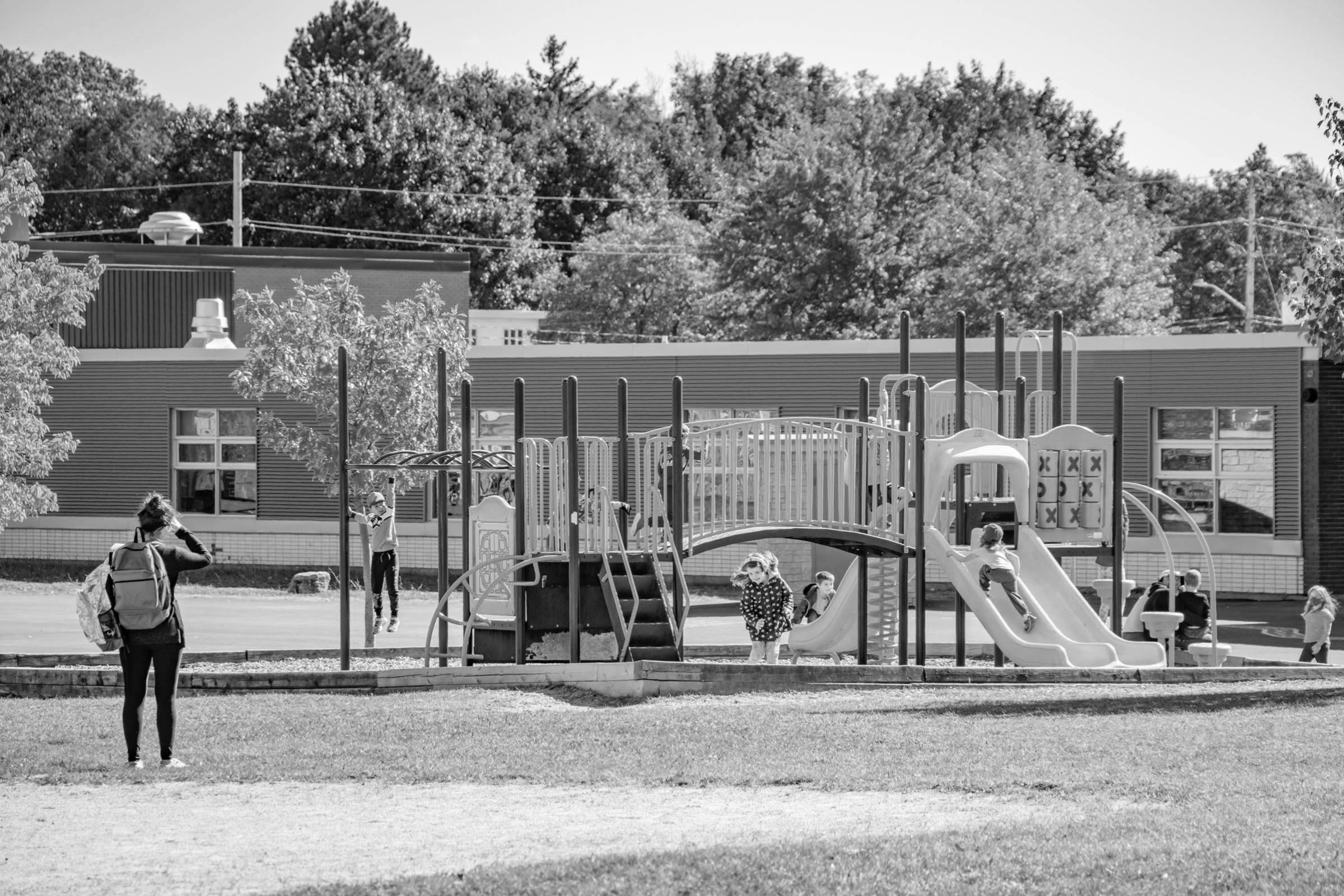 a photo of children playing at a playground while a parent wearing a backpack watches